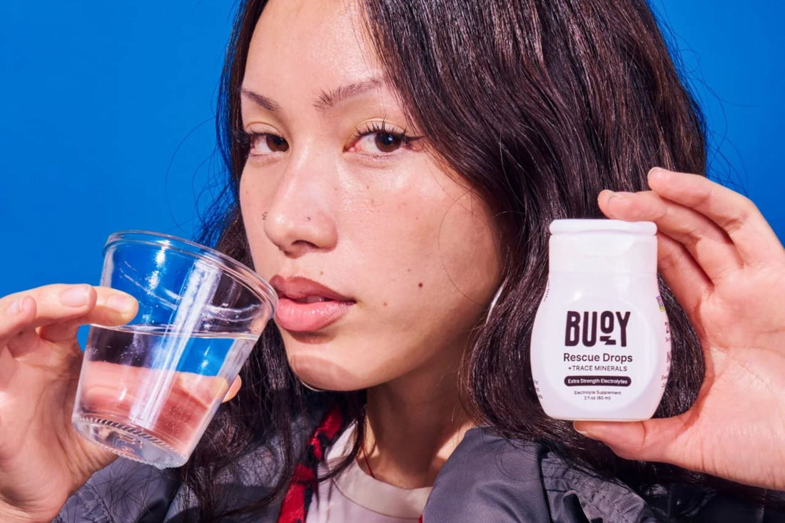 A woman holding a glass of water and a bottle of Buoy Rescue Drops electrolyte supplement, demonstrating proper hydration techniques for managing POTS symptoms.