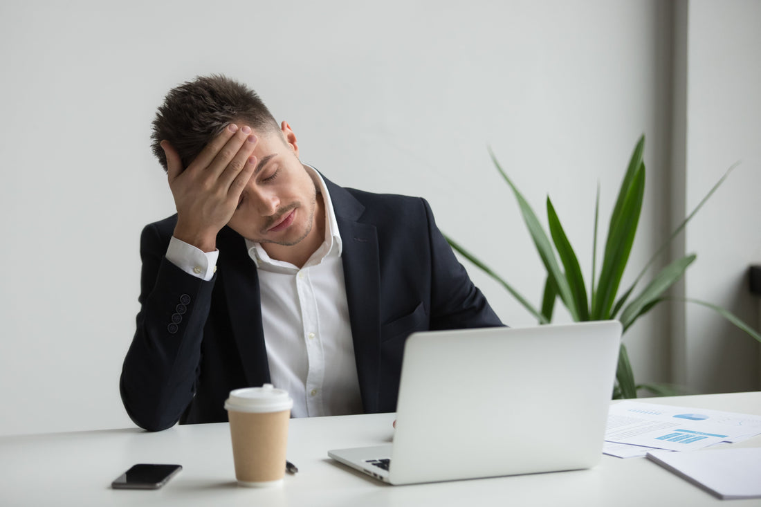 Stressed businessman in a suit sitting at his desk with eyes closed and hand on forehead, feeling fatigued while working on a laptop.