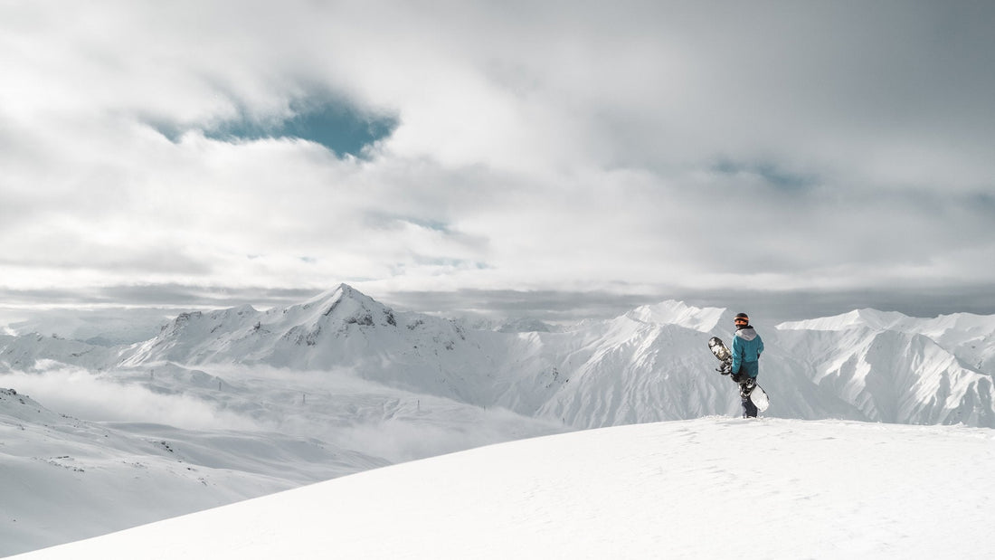 A snowboarder standing on a snowy mountain summit, gazing out over a vast, cloud‑shrouded alpine landscape.