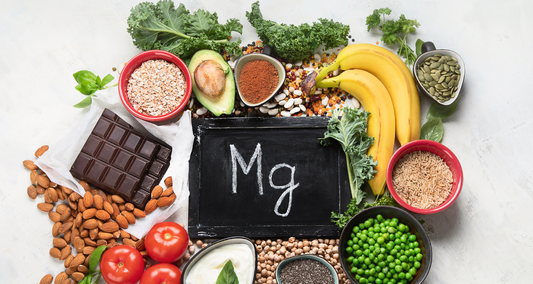 A chalkboard labeled “Mg” surrounded by magnesium‑rich foods almonds, dark chocolate, bananas, avocado, kale, pumpkin seeds, peas, tomatoes, oats, rice, and yogurt, arranged on a light surface.