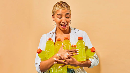 Smiling woman holding several bottles of bright yellow sports drinks, appearing surprised and delighted against an orange background.
