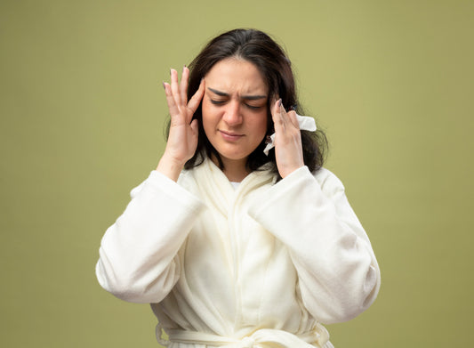Woman experiencing migraine pain holding temples with eyes closed on neutral background.