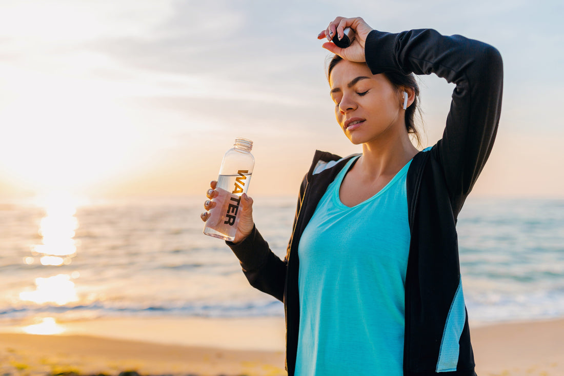 Woman hydrating with water bottle after exercise on beach at sunrise, supporting hydration routine for migraines and electrolyte balance