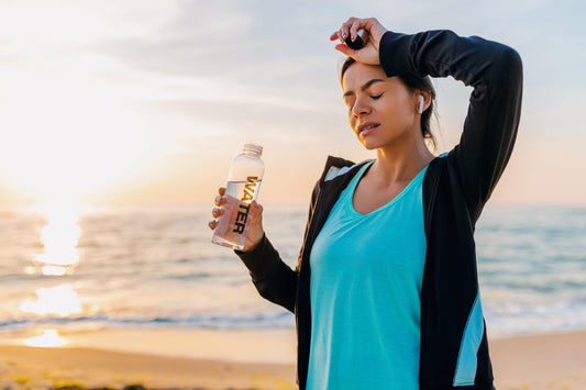 Woman hydrating with water bottle after exercise on beach at sunrise, supporting hydration routine for migraines and electrolyte balance