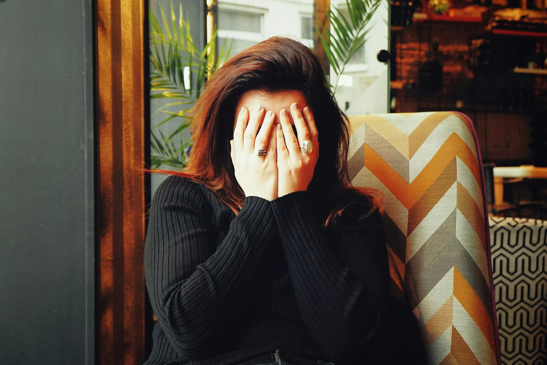 Person sitting indoors covering their face with both hands, appearing stressed or overwhelmed while seated in a chair near a window.