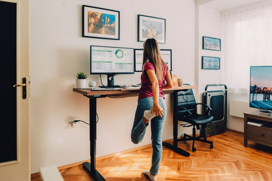 A woman stands at a standing desk, shifting her weight and stretching her leg while working to prevent triggering her POTS symptoms from prolonged standing.