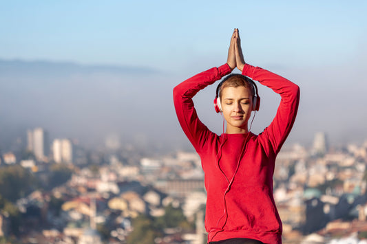 Young woman meditating with headphones outdoors, eyes closed, practicing mindfulness and mental clarity against a blurred cityscape background.